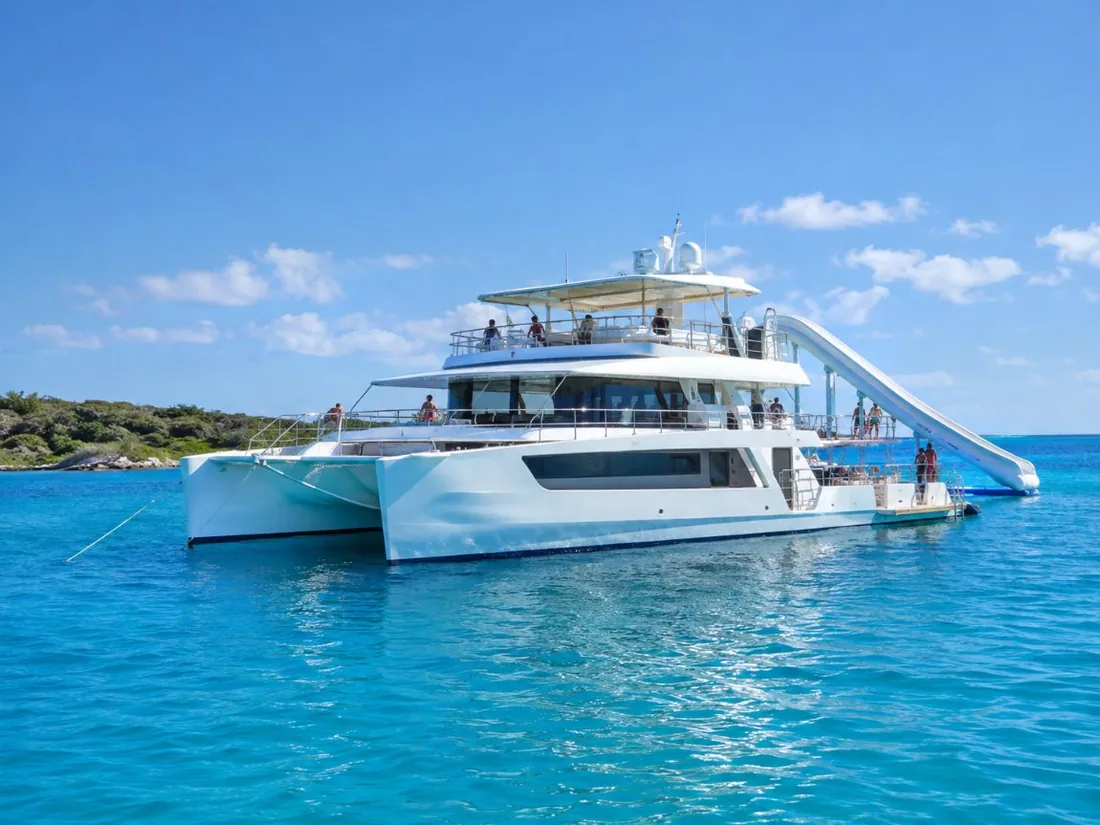 A large white catamaran with a water slide off its upper deck on calm blue water