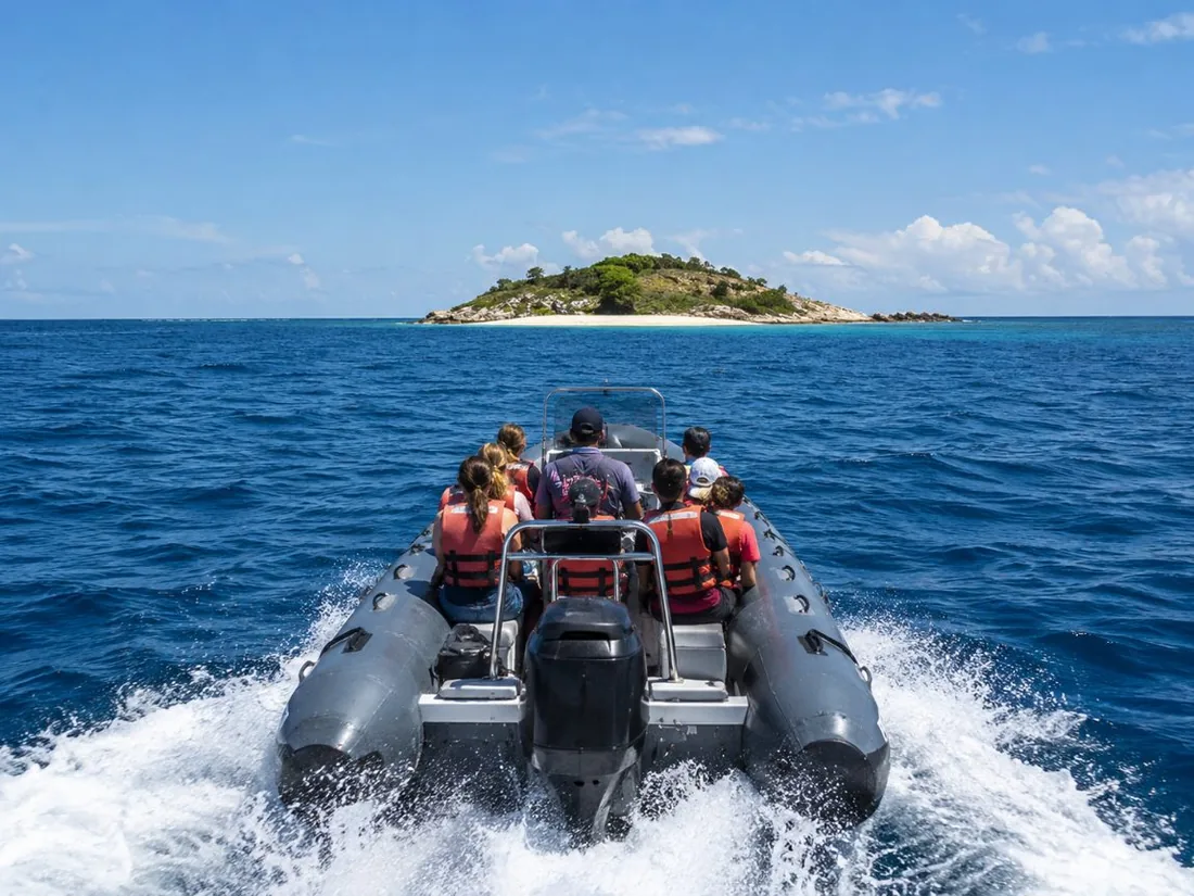 A fast rigid inflatable raft with passengers crossing open blue water toward a small crescent islet